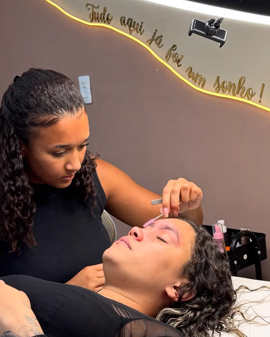 Aluna do curso fazendo design de sobrancelhas em uma modelo, no studio, sob o letreiro “Tudo aqui já foi um sonho!”